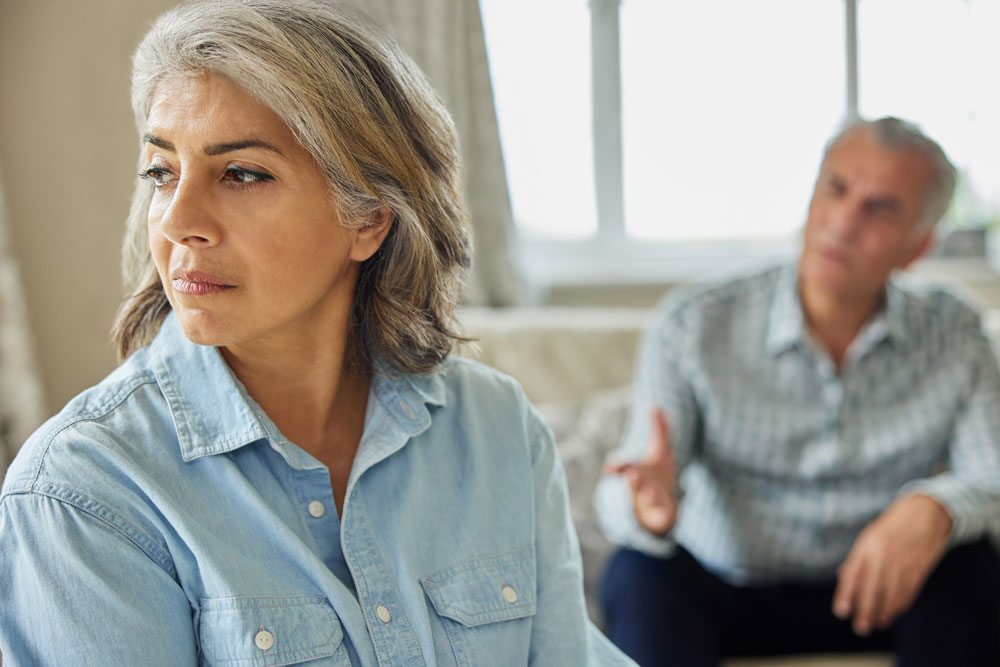 An older woman in a blue button down looks away from a man who is out of focus and speaking to her. 